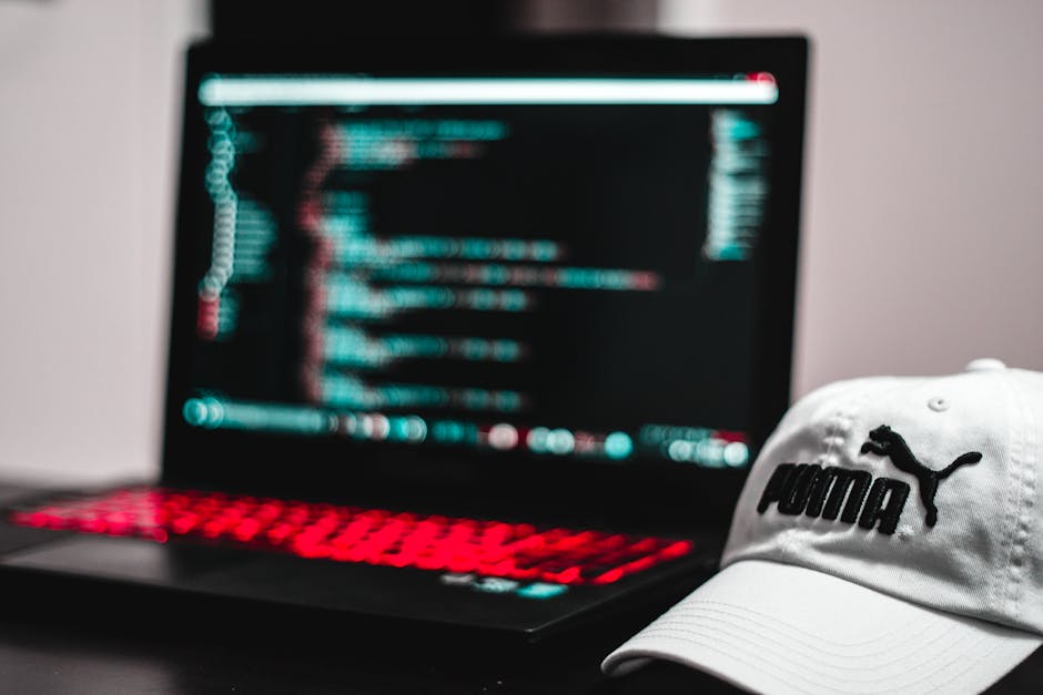 A close-up view of an open laptop with a red-backlit keyboard displaying lines of code on a dark screen, positioned on a surface inside a property. To the right of the laptop, there is a white Puma-br