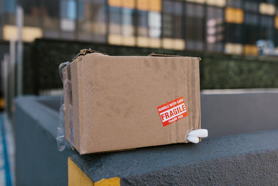 A small cardboard box with a red and white 'Fragile' label and a 'Handle With Care' sticker is positioned on the edge of a dark, textured surface outside a residential building. The box is sealed with