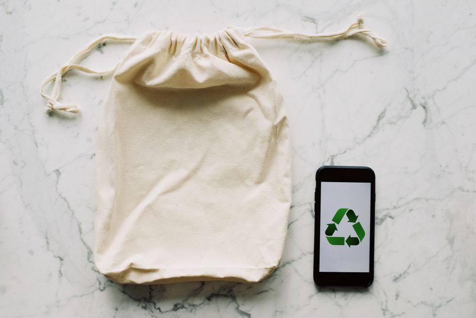A cream-colored cotton drawstring bag placed on a white marble surface, alongside a black smartphone displaying a green recycling symbol on its screen. The bag appears to be used for packing or storin